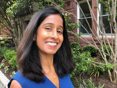 Qudsiya is wearing a blue dress and standing beside plants in front of a building on a city street.