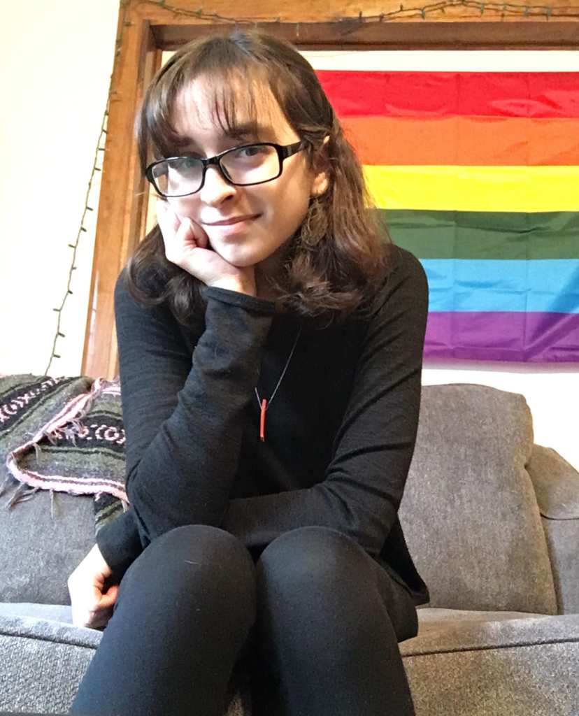 Rachel, a white femme-presenting person sits in front of a pride flag. She has brown shoulder-length hair, and is wearing bronze earrings, black clothing, and black rimmed glasses. A little red piece of wood dangles from a thin silver necklace chain around her neck. She is sitting on a grey couch and rests her chin in one hand with one elbow on her knee. She grins slightly at the camera.