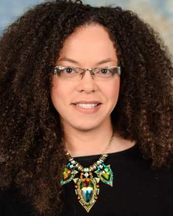 Headshot of a light complected African American woman, late thirties, with curly brown hair and glasses. She is wearing a black top and a large statement necklace.