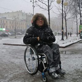 A middle-aged white man wearing eye glasses. He is sitting in a manual chair outdoors on a gray snowy day. He has on a winter jacket with a furry hood. His hands are stuffed in his pockets. The background is an urban setting with buildings, trees and sidewalks.