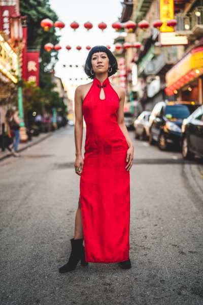 Rochelle Kwan, a Chinese-American woman with short black hair, stands in the middle of a San Francisco Chinatown street looking down at the camera with a soft smile. She wears a long, red cheongsam dress, black boots, gold hoop earrings, and a jade ring. In the background are rows of hanging red lanterns, neon signs with Chinese characters, and parked cars. This photo was taken by her sister, Elodie Kwan.