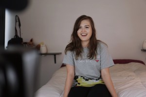 Image Description: Rikki in the middle of filming. She is sitting on a bed in a bedroom, talking/smiling at the camera. Photo credit: createitkind.org