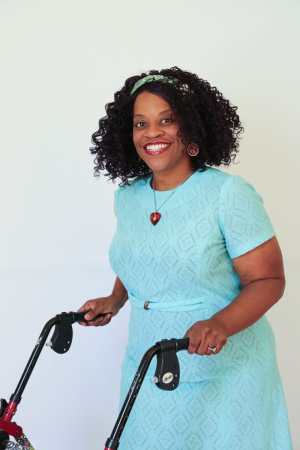 Robin, a brown woman, is posed and smiling. Wearing a light blue-green lace vintage dress, she is holding the handles of her walker.