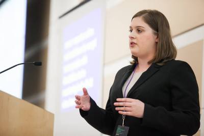 Profile of a young white woman with shoulder-length brown hair and a black blazer. She is speaking in front of a lectern. Behind her is a screen projected with a slide.