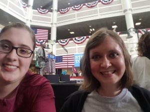 Selfie of two young white women in a large call. Behind them you can see a stage with a lectern, multiple American flags hung up and patriotic bunting on multiple floors.