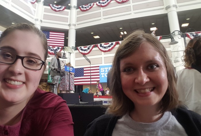 Selfie of two young white women in a large call. Behind them you can see a stage with a lectern, multiple American flags hung up and patriotic bunting on multiple floors.