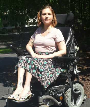 A young, white woman with short blonde hair is pictured outside. She is wearing a pink tee-shirt, black skirt with pink and green design, and matching heels. She is smiling toward the camera and seated in a black power wheelchair.