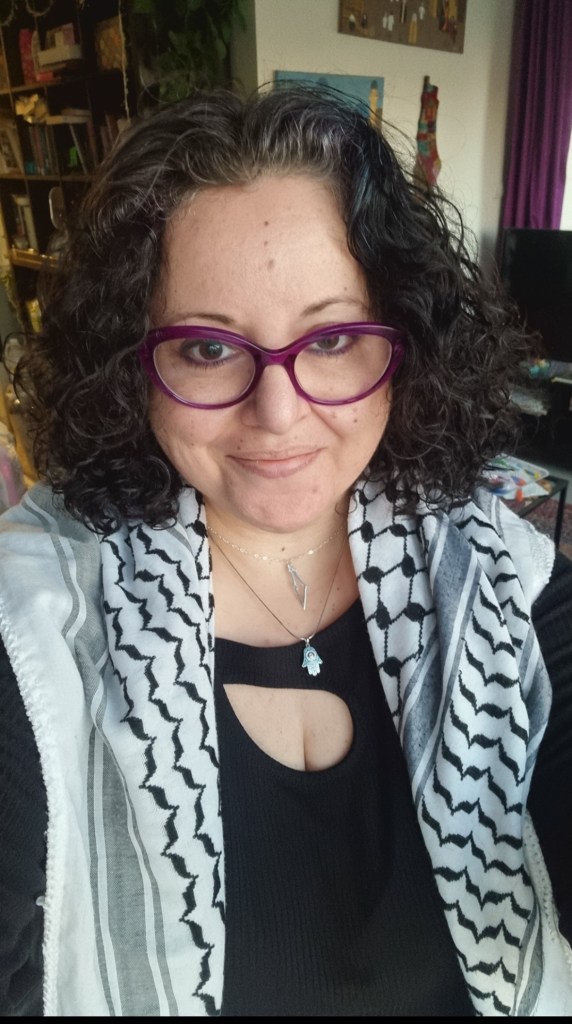 A Palestinian Jordanian woman in her 40s with dark curly hair, purple glasses wearing her Palestinian kuffiyeh over her shoulders as well as a necklace of the map of Palestine, behind her on the wall is an embroidered map of Palestine