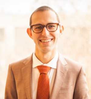 Sean Betouliere – a white man in his mid-30s with close-cut hair, thick-framed glasses, a brown suit, and an orange tie—smiles for the camera