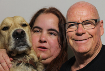 A photo featuring Maia Scott, Ron Jones, and Maia's guide dog was taken on August 14, 2014: close up portrait of a guide dog, a woman and man all sitting next to each other looking at the camera. The guide dog on the far left is a golden retriever with its nose and face pointed at the camera. The woman in the middle appears to be white and has long brown hair. The man on the right appears to be white, is bald, wearing glasses, dressed in a black shirt, and smiling at the camera.