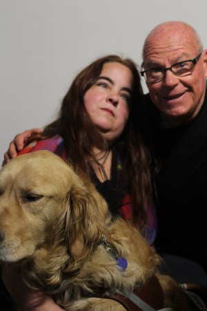 Photo of two people against a blank white background. An older white man with glasses has his arm around a young white woman with long brown hair. The man is wearing a black top and the woman is wearing a multi-colored top. In front of the woman is a golden retriever, her service animal.
