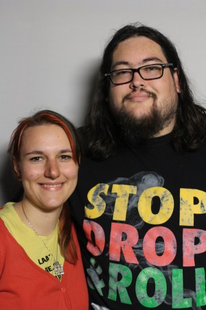 Photo of two people against a blank white background. On the right is a tall young man with long dark brown hair, a beard and glasses. He is wearing a black t-shirt that says “Stop, Drop, & Roll” in yellow, red, and green for each word. Next to him on the left of the image is a young white woman with long brown hair and streaks of red highlights. She is wearing a red hoodie with a yellow t-shirt. Both are smiling at the camera.