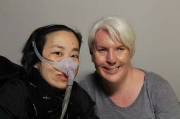 Photo of two people against a blank white background. A white woman on the right has white-blonde hair pulled back. She is wearing a scoop neck gray top. On the left side is an Asian American woman in a wheelchair wearing a black jacket and black patterned scarf. She is also wearing a mask around her nose with a tube. Both are smiling at the camera.
