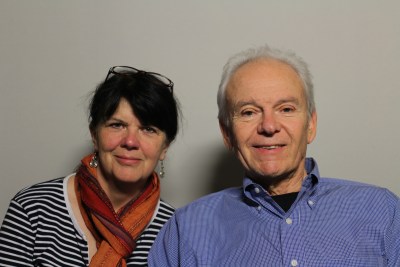 Photo of two people against a blank white background. An older white man is on the right side of the image. He has short white hair and wearing a button-down long-sleeved blue shirt. To the left of him in the image a white woman with dark brown hair. A pair of glasses are positioned on top of her head. She is wearing dangly earrings, a multicolored scarf wrapped around her neck and a black-and-white striped shirt. Both are smiling at the camera.
