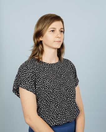 In this portrait, I’m facing askew from the camera and smiling slightly. I have medium-length wavy blonde hair, and I'm white. I'm 30, but people often tell me I look younger. I’m wearing a shirt with a pattern of white squiggles on black paired with bright blue dress pants.