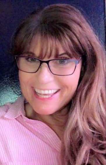 A mixed Native and white person with large square glasses and long, flowing brown hair, looking directly at the camera and smiling widely and warmly. They are wearing a light pink collared shirt.