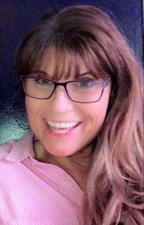 A headshot showing Sharon with long brown hair, wearing large square glasses, looking directly at the camera and smiling. They are wearing a light pink collared shirt.