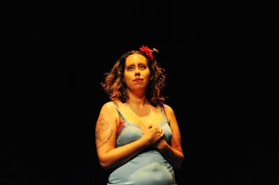 A woman of color with curly brown hair on stage (the background is all black). She has a red flower in her hair. Her hands are clasped together at her chest. She is wearing a light blue tank top.