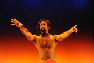 Image of a Black man bare-chested on a stage. His hands are wide open and outstretched. He has a beard and short dreadlocks. The stage is light in orange, black and blue starting from the floor up.