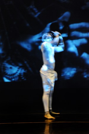 Image of a person dressed in white on stage. The person's arms are raised. A projected image is projected over the person's body in different shades blue.