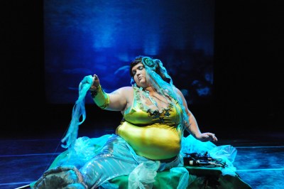Large woman sitting on a stage dressed in bright colors aqua colored fabric flows over her legs, she has aqua hair extensions as well. She looks like a dreamy sea creature. Her top is a form-fitting tank style in bright citron yellow-green. Behind her are blue lights on the stage.