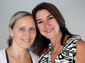 Image of two women smiling at the camera standing close to each other. On the left is a woman with blonde hair and blue eyes and on the right is a woman, her mother, with long brown hair and brown eyes