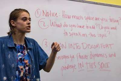 A white femme-presenting person with dark blonde hair stands in front of a white board with a blue floral shirt and a jean jacket. The white board has red text on it, which states: “1. Notice: How much space are you taking? 2. What do you have to add? Do you need to speak on this topic? 3. Lean into discomfort. 4. Recognize power dynamics and privilege in this space.”