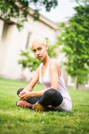 A light-skinned Black woman with a blonde buzzcut and a lavender dress sits on the grass in front of two Brown University buildings, one of which was built by enslaved Black people. She is looking into the camera.