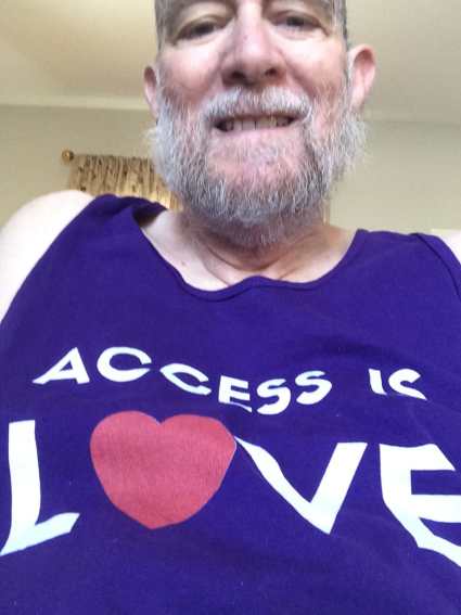 Steve Brown, an older, smiling, man with graying brown beard is shown in his office in Arizona, wearing a sleeveless, “Access is Love” purple tank top.