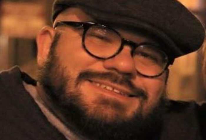 White, cis man with dwarfism smiles at the camera. He has a shaved head and short beard andis wearing dark rimmed glasses, a dark driver's cap, and dark cardigan over a light t-shirt.