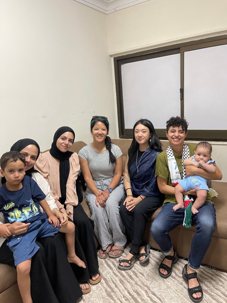 In a white-walled corner of a room with windows where the blinds are drawn, a few members of the Sulaiman family, from the left - Nada with black hijab, white blouse, and long black skirt holding her four year old son; her sister Wafaa in a beige-pink blouse and black hijab and long black skirt; comrade Daranee with long black hair in a ponytail that falls to her right, gray blouse and striped pants; and comrade Joie with long black hair, blue shawl, and black pants. dena is to the furthest right and is holding the infant; they have curly black hair, green shirt, keffiyeh scarf and blue jeans. They all sit on a brown couch and everyone is smiling except for the children. 
