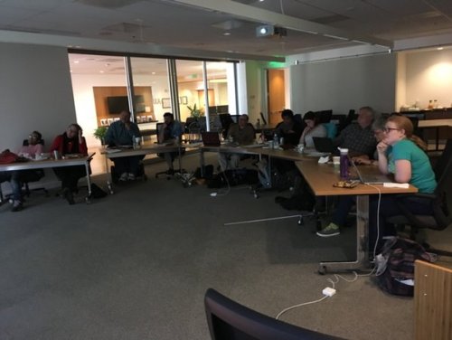 Photo of a darkened room with a row of tables facing a screen. Many people are sitting at tables looking intently at something.