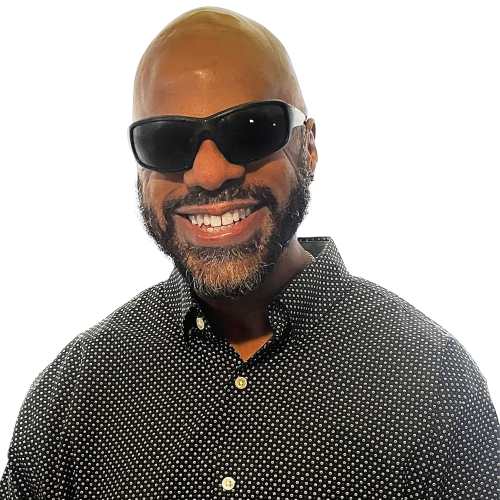 Photo of Thomas Reid, a brown skin Black man with a clean-shaven bald head and full neat beard,  smiles into the camera. He wears dark shades and a black and white checkered button up shirt.
