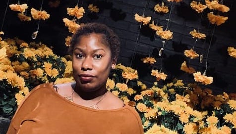 Teefloetry is standing behind a black brick-coated wall with the flowers hanging. She is posed towards the camera with short black & brown mixed curls.