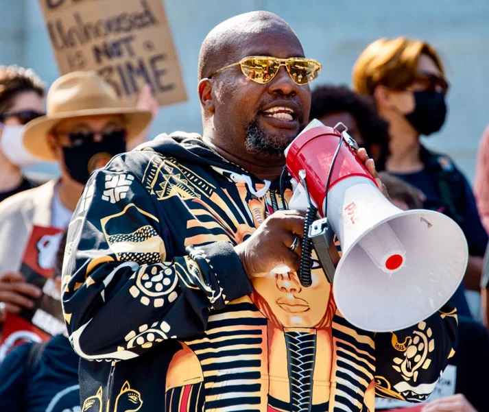 Theo Henderson, a Black man with a shaved head, goatee, gold-tinted sunglasses, and wearing a hoodie with an image of Tutankhamun on it, speaks into a red and white megaphone at a protest. Behind Theo is a crowd of people wearing face masks, with one holding up a sign that says "Being unhoused is not a crime." Credit: WeTheUnhoused.com