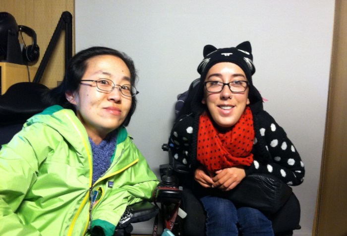 Image of two women with disabilities who use wheelchairs. On the left is an Asian woman with glasses and a green jacket. On the right is a white woman with glasses and a black sweater with white polka dots and a red scarf