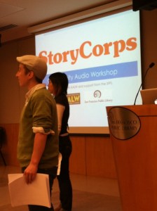 Two people standing profile facing the left side of a room, in the backdrop is a projector screen with the words, "StoryCorps" in large orange letters