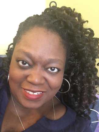 A Black woman with long curly hair smiling at the camera. She is wearing a navy shirt, a silver necklace and silver hoop earrings.