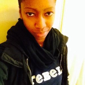 TL , a black person with short hair takes a selfie while wearing a black hoodie & black scarf with a navy shirt that has white words "REBEL."