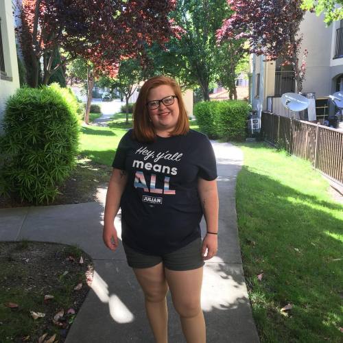 A standing picture of Tory Cross, a white woman with red hair and black glasses, smiling and wearing green shorts a blue shirt that says “Hey Ya’ll Means All” with hey y’all means in white font and All in stripes of blue, pink and white. There is a sidewalk and greenery behind her.