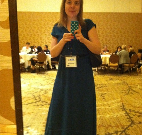 Image of a white woman taking a selfie of herself in front of a mirror. She is wearing a long blue dress and holding her cell phone in front of a mirror.