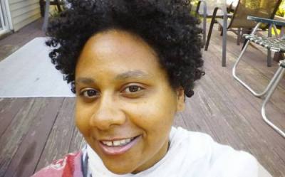 Black woman with curly natural hair wearing a white top with streaks of pink, red, and gray. She is smiling at the camera.
