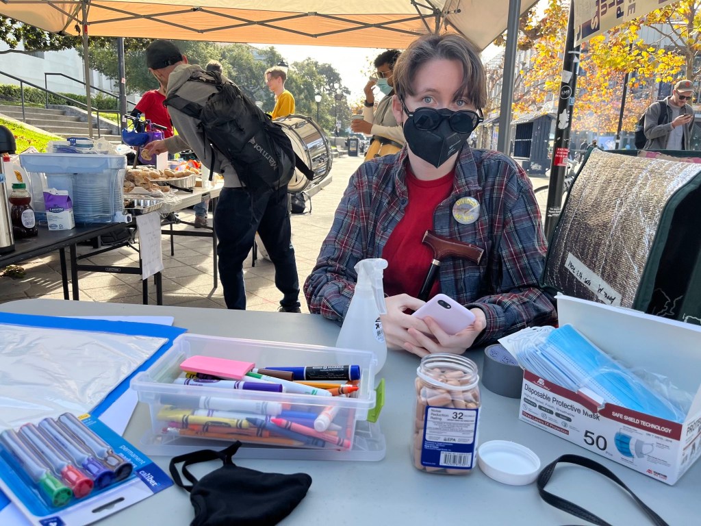 Disabled UC Grad Worker & UAW union member Ros wearing shades, an N95, plaid shirt with union red beneath it and their cane leaned against them with their cell phone in their hand. They are sitting down in front of a table with free ear plugs, free masks, and pens. Behind them is free food and other masked union members. Photo of UC Berkeley Accessible Mutual Aid Tent and union member Ros by Pratiti Ketoki. 2022. Used with permission.