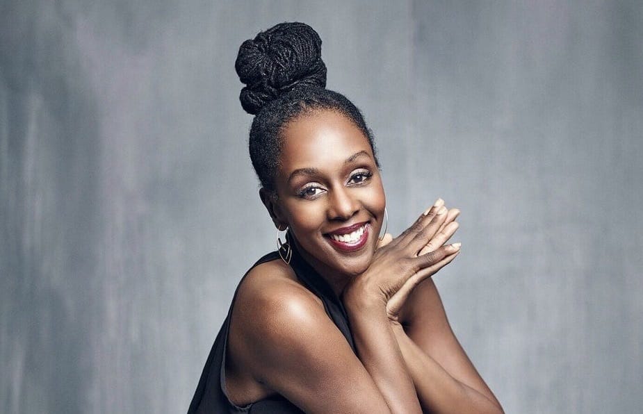 Stephanie Thomas, a Black woman with oval shaped eyes, round nose, smiling. Her chin is resting on her clasped hands. Her hair locs are pulled up in a high bun and wearing a sleeveless black top. The background is a grayish black backdrop. My condition’s name is Symbrachydactyly. I am a congenital amputee missing digits on my right hand and feet. Photo credit: Benjo Arwas @benjoarwas