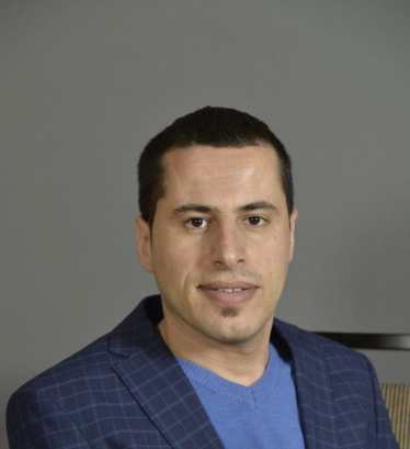 Photo of Mustafa Rfat, a man with short dark brown hair wearing a navy blue blazer with v-neck blue shirt. Behind him is a dark gray background.