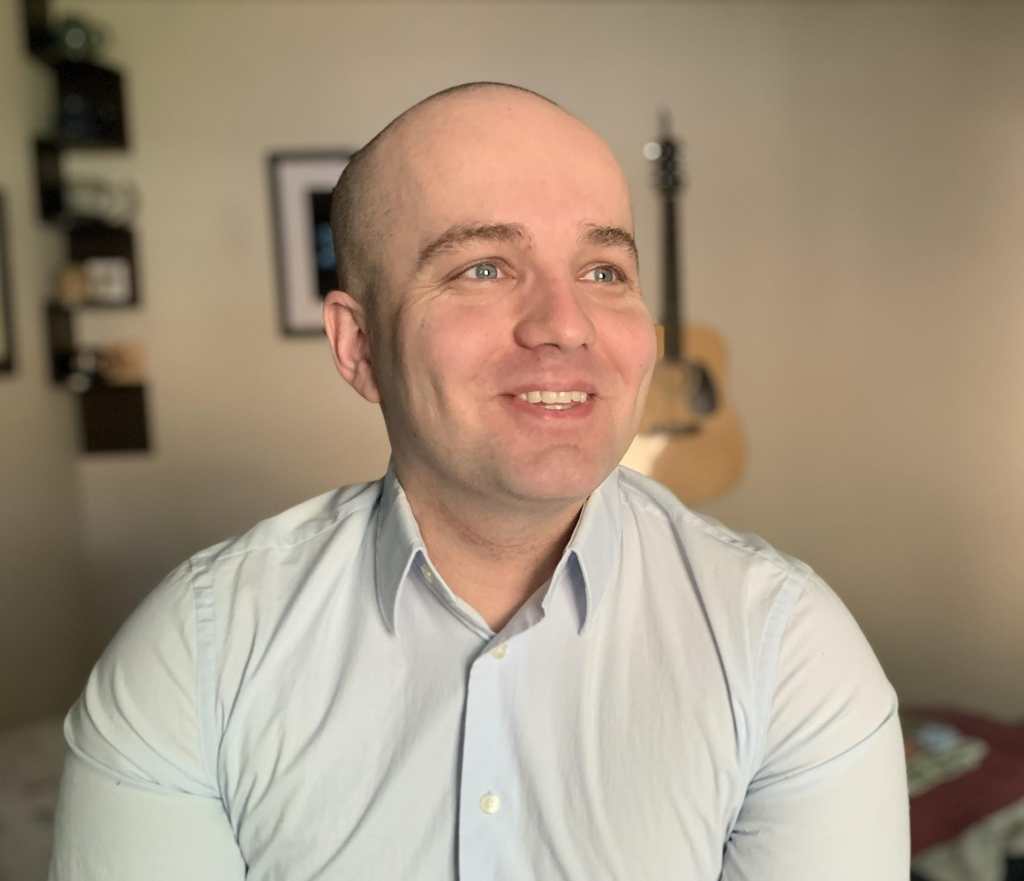 A photo of Jamison Hill, a young white man with an almost clean-shaven head with expressive blue eyes. He is looking away from the camera, slightly to the right and wearing a light blue, long-sleeved button down shirt. He is smiling.