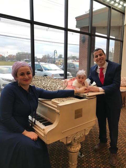 Photo of three people gathered around a white grand piano. On the left sitting on the piano bench, Nazli Khazirova, a woman with a purple head covering and a navy blue outfit smiling widely. Her arm is extended toward Mustafa Rfat on the right, a man wearing a navy blue business suit with a white shirt and red tie who is also smiling. In the center, laying on top of the piano is Defne Rfat, a little baby girl with a white flower-like headband and a white and pink tulle outfit like a ballerina