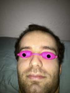 A photo of Jamison Hill, a young white man with short brown hair and a half-grown beard. He is lying in bed wearing hot pink tanning goggles over his eyes that aid him in his writing.