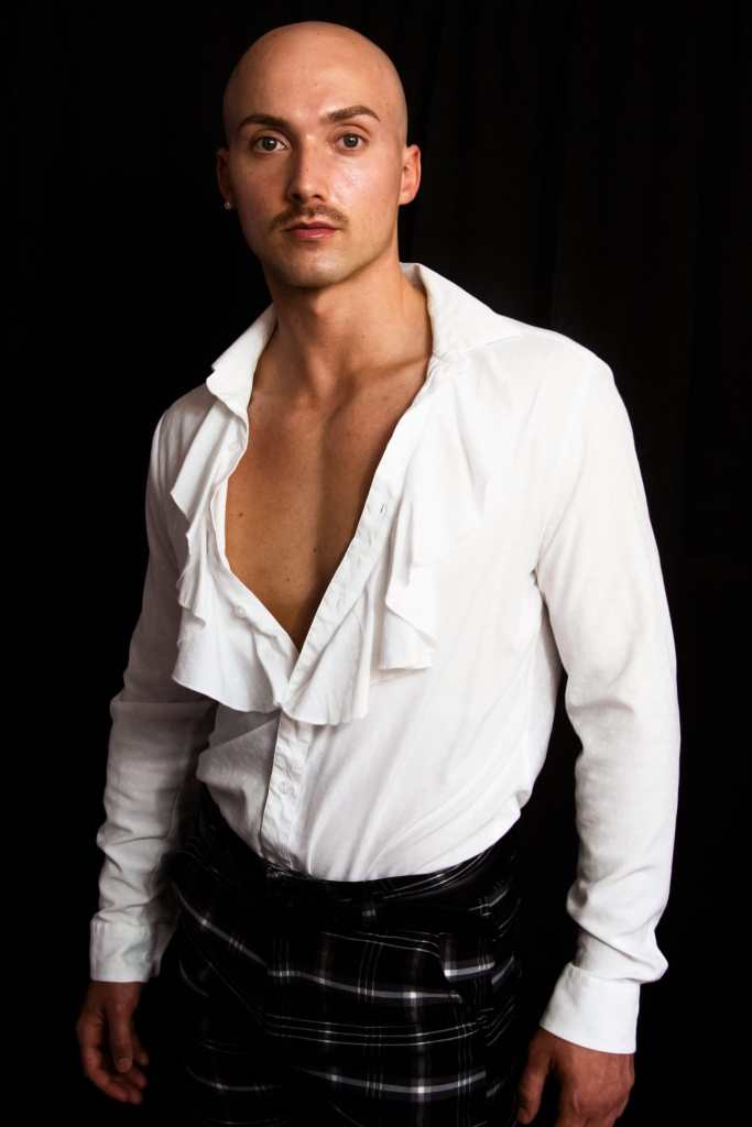 J. Logan Smilges, a white gender nonconforming trans person with a mustache, looks toward the camera without smiling. They are wearing a long-sleeve white shirt with ruffles down the middle and black pants with a checkered print. Photo credit: Darius Stokes