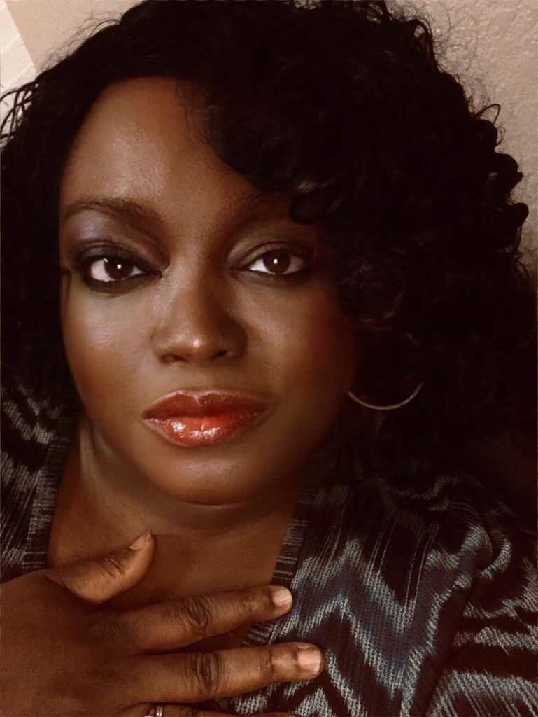 A head and shoulders picture of Tinu Abayomi-Paul, against a light brown walk. She is a dark skinned woman with chin length curly black hair. She has on red lipstick and her left gold colored hoop earring is visible. She is not smiling, but looks content. She has a hand lightly pressed against her chest and is wearing a dark blue, light blue and black blouse patterned in zigzag stripes. She's looking directly into the camera.